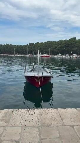 A small red and white boat is moored in calm water, facing the camera. The boat has a metal railing around its front and a single mooring line connecting it to an unseen anchor. Its reflection is visible in the water. In the background, a line of trees forms a dense green forest along the shore. Further back, several sailboats with masts are anchored in the distance, under a partly cloudy blue sky. The foreground features a stone walkway or dock.