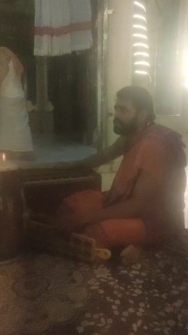 A person wearing an orange garment sits on a patterned rug and plays a drum. They hold a stick in their right hand and strike a drum with it. A second drum is placed in front of them, and it also appears to be struck by the person. There are shelves with white fabric rolls in the background. The scene is dimly lit, with some light sources reflecting off a surface.