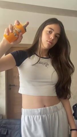 A young woman with long brown hair sprays perfume on herself. She is wearing a white crop top with blue sleeves and grey sweatpants. The crop top has the word "CHICAGO" printed on it. She is standing in what appears to be a bedroom, with a closed wooden door visible in the background. She sprays the perfume once, then purses her lips. She appears to be looking at the camera. The perfume bottle is orange with gold accents.