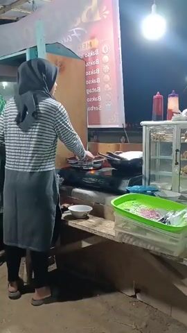 A woman wearing a black hijab and a striped shirt is cooking skewers over a gas stove. She is standing in front of a food stall at night. There is a sign behind her with text and images. In front of her, there is a pan, a bowl, and a green container. The background is dark, suggesting it is nighttime.