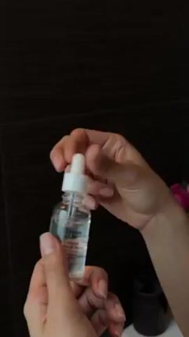 A pair of hands is seen holding a small, clear glass bottle filled with a transparent liquid. The bottle is labeled "Smooth & Lift Collagen Facial Serum." The right hand unscrews a white dropper from the bottle. The dropper is used to extract a small amount of the serum from the bottle. The background is a dark tiled wall. Beside the bottle is a white sink and a dark container. In the background are some pink flowers.