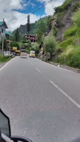 The video shows a view from a moving vehicle on a paved road. To the left, there is a sidewalk with parked cars and a yellow sign. Further up the road, a white and red ambulance is visible. Beyond the ambulance, a large truck is driving. On the right side of the road, a steep, rocky embankment covered in green foliage rises sharply. In the background, a village or town is nestled on the hillside, with houses and buildings visible among the trees. The sky is blue with scattered white clouds. The overall scene depicts a scenic mountain road with traffic.