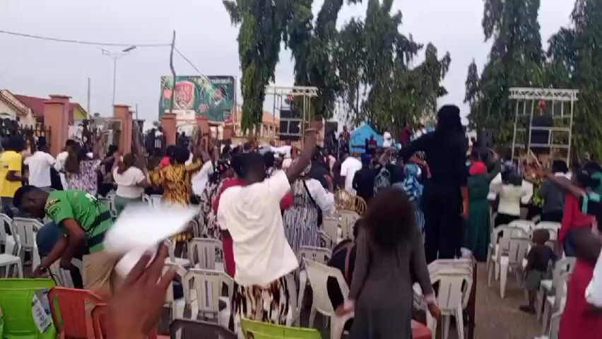 The video captures an outdoor gathering of people, possibly a religious service or a public event. A large crowd is visible, with many individuals standing and some seated in rows of plastic chairs. The atmosphere appears lively, with people raising their hands and some dancing. A stage or platform with sound equipment is set up in the background, suggesting a performance or speech is taking place. In the distance, there are trees, buildings, and a large billboard. The lighting suggests it is daytime, possibly overcast or early morning/late afternoon.