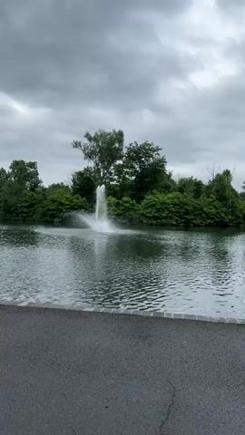 A wide-angle shot of a park on a cloudy day. In the foreground, a paved pathway runs along the bottom of the frame. Beyond the pathway is a lake with gentle ripples on its surface. A fountain in the middle of the lake sprays water upwards. Lush green trees and foliage line the far bank of the lake. In the middle distance, a modern building and a smaller, darker building with a sloped roof are visible amongst the trees. The sky is overcast with heavy grey clouds.