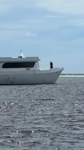 The video shows a large, white boat with two visible people on deck, floating in a sea or ocean under a partly cloudy sky. The camera appears to be on another boat, initially focusing on the first boat's front and then shifting slightly to reveal a portion of the camera boat's frame. The water has a slightly choppy surface, and a light-colored sandbar or beach is visible in the distance. A small, blue boat is attached to the back of the white boat.