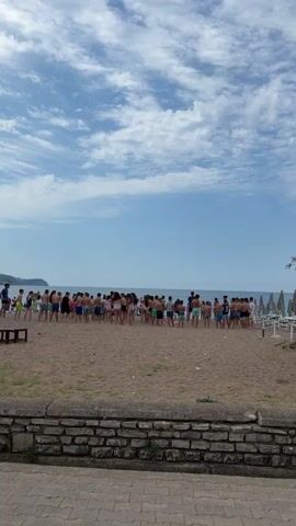 The video captures a large group of people, predominantly children and young adults, gathered on a sandy beach in Bečići, Montenegro. They are mostly dressed in swimwear and are facing the calm, blue sea. The group appears to be participating in an organized activity or game, with some individuals raising their arms and moving dynamically, suggesting excitement or engagement.

The weather is bright and sunny, with a clear blue sky featuring scattered white clouds. The setting is a typical resort beach, indicated by rows of folded beach umbrellas and sunbeds visible on the right side of the frame, along with a palm tree. A low stone wall and a paved pathway are in the immediate foreground, separating the viewer's perspective from the beach itself. The time of day appears to be daytime, likely morning or early afternoon, given the bright illumination. No discernible text is visible in the frame. The overall scene depicts a lively and active gathering at the beach.