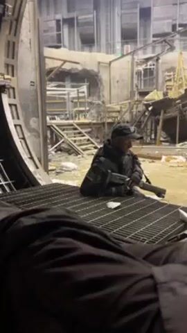 The video shows a man in a black uniform and cap holding a shotgun while sitting on a metal grate in a war-torn environment. The man has a Russian flag patch on his sleeve. The background depicts a dilapidated building with debris scattered throughout.