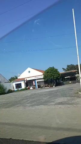 The video shows a view from inside a vehicle, likely a car, driving down a road on a clear day. The camera pans across a scene that includes several buildings, a parking area, and some greenery.

In the first segment, a small, white building with a blue trim is visible. It appears to be a business, possibly a shop or cafe, with a sign that reads "RESTAURANTE." There's a paved area in front of it.

The next segment shows a collection of large, colored trash bins, including blue, silver, and green ones, situated near some construction materials and a utility pole with traffic lights.

Then, a red building with white trim and a covered outdoor area is seen, with trees and foliage in front. There's a gate with a "no parking" sign.

Following that, the view shifts to a scene with a truck covered by a striped awning, parked in front of a building. Another awning is visible to the right. The truck has a license plate.

The final segment shows a row of vehicles parked in front of a building with an orange tiled roof. The building has signs with text, including "PRÍNCIPE DO ISAIAS." A silver minivan, a white van, and other cars are parked in a paved area in front of the building. The view includes a side mirror of the vehicle from which the video is recorded.