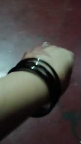 The video shows a close-up view of a person's arm and wrist with several black bangles. The arm is positioned against a dark reddish-brown surface, likely a floor. The person's hand is blurred and appears to be moving, possibly shaking the arm or engaging in an action that causes the bangles to move. The lighting is dim, creating shadows and highlighting the shininess of the bangles.