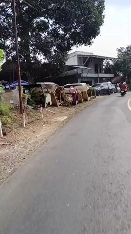 The video shows a street scene with several abandoned car bodies lined up along the side of the road. The car bodies appear to be old and rusted, some are yellow and others are a darker color. There are also some functional vehicles parked further down the road, including a black pickup truck, a white van, and a white car. A red motorcycle is also visible. In the background, there are buildings, trees, and a mosque with a green dome. The road itself is paved with asphalt and has a yellow line marking the lane. There are people visible riding motorcycles in the distance. The scene appears to be in a rural or semi-rural area.