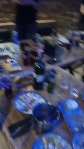 The video shows a restaurant with multiple tables and chairs, some of which are occupied by people. At the beginning, the focus is on a table with food, drinks, and empty dishes, suggesting a meal has been consumed. The scene then transitions to a busier area with more people, some dancing and raising their arms, possibly in response to live music or a DJ. The lighting changes frequently, with pink, green, and blue hues illuminating the room and the patrons. Throughout the video, the camera movement is shaky and unfocused, making it difficult to discern details. The atmosphere appears lively and social, indicating a festive environment.