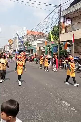 The video shows a street parade. Several people are walking down the street, dressed in colorful traditional costumes. The costumes are elaborate and feature gold embellishments, feathered headdresses, and brightly colored fabrics. Some of the participants are wearing black leggings and white sneakers. The parade appears to be taking place on a sunny day, with buildings lining the street and power lines overhead. There are spectators on the sidewalks watching the procession. Some of the spectators are holding umbrellas to shield themselves from the sun. The overall atmosphere seems festive and celebratory.