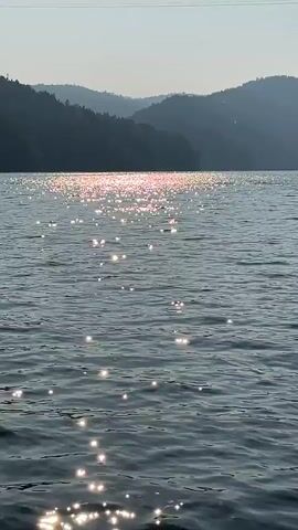 The video shows a body of water with shimmering sunlight reflecting off its surface. In the background, there are hazy, silhouetted hills. The camera appears to be moving slightly, possibly on a boat, as the water ripples and the light sparkles shift. No people or specific objects are clearly visible. The overall atmosphere is calm and serene.