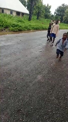 Children are running down a wet, gray asphalt road. In the background, there are green trees and bushes, and some buildings. The children appear to be of various ages and are wearing casual clothing and school uniforms. The video is shot from a low angle, following the children as they run.