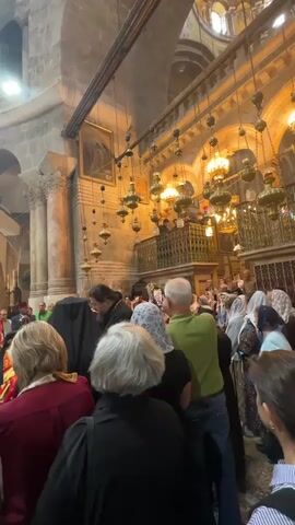 The video shows a crowd of people inside a large, ornate church. The interior is grand with stone columns, arches, and a balcony. Above, a multitude of hanging lamps illuminate the scene. A religious service appears to be in progress, with priests in traditional robes near the front of the crowd. Some members of the crowd are taking photos or videos with their phones. The atmosphere is solemn and reverent.