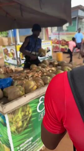 The video shows a man cutting into coconuts at a roadside stand. He is wearing a dark blue vest and a black beanie. He stands behind a table covered with coconuts, many already partially cut open. He uses a large knife to cut open each coconut, skillfully creating a hole to access the coconut water. The stand has signs displaying food offerings and contact information. A person wearing a red shirt is partially visible in the foreground. In the background, people walk by and other food stands are visible. The scene appears to be set outdoors in a bustling commercial area.