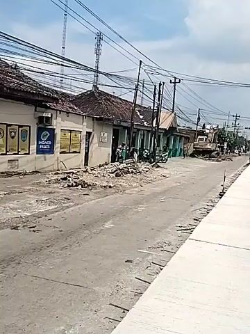 The video shows a street scene with a large excavator parked on the side of the road, surrounded by debris and rubble. Several people are walking on the sidewalk and along the road, and a few motorbikes and a large truck are driving past. The buildings on the side of the road appear to be old and somewhat dilapidated, with some of them showing signs of disrepair. Overhead, a dense network of power lines and telephone wires crisscrosses the sky, adding to the cluttered visual environment. The sky is partly cloudy, with patches of blue visible between the clouds.