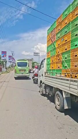 A white truck carrying a tall stack of green and orange plastic crates drives on a paved road under a partly cloudy sky. The crates appear to be poultry crates. The truck is in the right lane of the road, which appears to be a two-lane road with a white dashed line separating the lanes. To the left of the truck, a green van is driving in the left lane, and several motorcycles are also in the left lane. A few people are visible on the motorcycles. In the background, there are trees and buildings, and utility poles with wires. The overall scene appears to be a rural or suburban road.