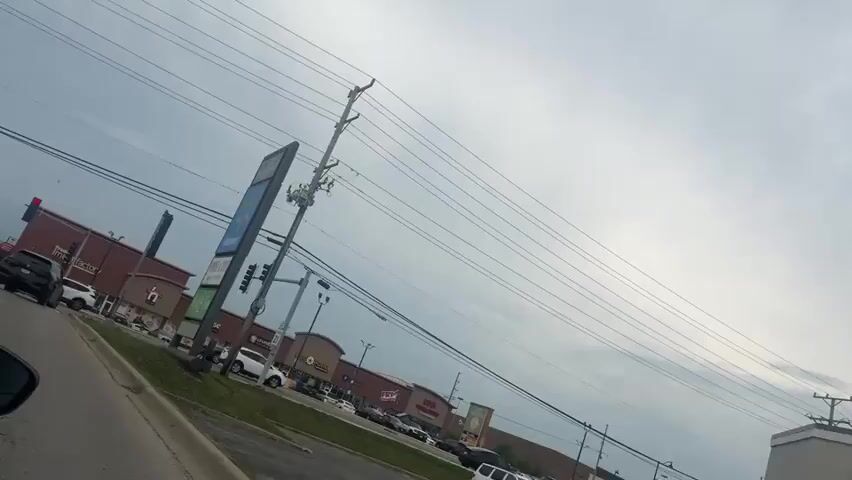 A dashboard camera records a view from a vehicle driving along a road. The sky is overcast with clouds. On the left side of the road is a sidewalk and then a grassy median. Beyond the median is a strip mall with several businesses, including a Walmart, a Smoke & Vape shop, and a business called "Do It For You Market." In front of the businesses is a parking lot with several cars parked and driving. A black SUV is in the foreground on the left, and further down the road, there are other cars. A large blue sign with the Walmart logo and other business names is visible. Power lines are strung across the sky, supported by utility poles. The camera is angled slightly to the right, giving a slanted view of the scene. The vehicle appears to be moving at a moderate speed.