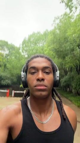 A young man with dark skin and long dark dreadlocks, wearing a black tank top, a silver chain necklace, and black over-ear headphones, is featured in this video. He is filming himself outdoors in what appears to be a residential area or park. The scene is characterized by lush green trees, well-maintained bushes, and paved pathways.

At the beginning, the man looks directly at the camera, then the view briefly pans down to show the wet paved path, green grass, and a barrier gate in the background, typically found at the entrance or exit of a residential complex. The camera then pans back up to the man, who is walking with a neutral expression. The video concludes with a quick upward tilt, showing the man's face followed by an out-of-focus tall building against an overcast sky.

The wet ground suggests it has recently rained or is still drizzling. The general environment points to an urban green space or a well-landscaped residential complex in Binjiang District, China, during daylight hours. No discernible text is visible in the video.