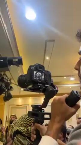 Here is a description of the video:
The video shows a Canon camera mounted on a gimbal, being held by a person. The camera screen displays a black and white image of a group of people, including one with a turban. The camera operator is standing in a room with gold accents and what appears to be an event space.  Other people are visible in the background.