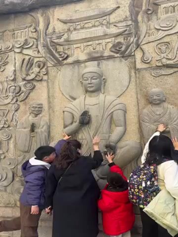In the video, several people are touching a stone relief carving of a seated figure with a halo, presumed to be Buddha, with other standing figures carved on either side. A pagoda is carved above the central figure. The people, including children and adults, reach out to touch the figure's hands and knees. The environment appears to be outdoors, possibly a temple or historical site.