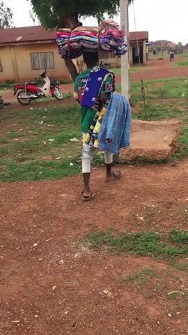 The video captures a person, likely a male vendor or hawker, walking away from the camera in Enyibichiri, Nigeria. The individual is heavily burdened, carrying a large, colorful bundle of fabrics or clothes balanced on his head, with more items draped over his shoulders and left arm. He is wearing a green t-shirt with a visible "Coca-Cola" logo on the back, light-colored pants, and open-toed sandals.

The setting is an outdoor, unpaved area characterized by reddish-brown soil and patches of green grass. In the background, there are simple, low-rise buildings, one featuring a red roof, and a wooden utility pole. A red and white motorbike is parked to the left of the frame.

The person walks towards a large, irregularly shaped stone or concrete block positioned in the mid-ground. He pauses next to the block and utilizes it to temporarily rest some of the merchandise he is carrying, appearing to adjust his load or take a brief respite. The scene unfolds during daytime, under what appears to be clear and bright weather conditions, depicting a moment of daily commerce in a local Nigerian community.