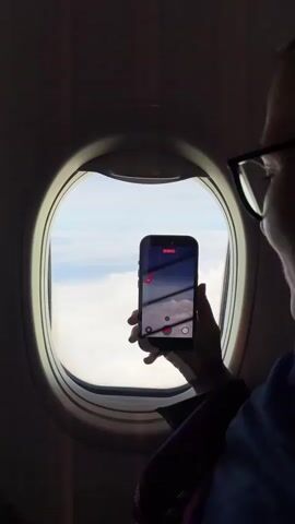 From inside an airplane, a person wearing glasses is filming out of the window with a phone. The screen of the phone shows the camera app, with a live video feed of the clouds outside the window and the plane's wing.