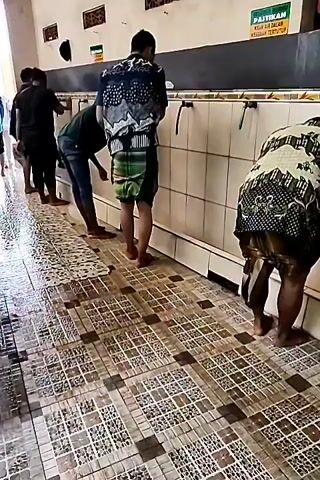 The video shows a group of men performing ablution, a ritual cleansing performed by Muslims before prayer, at a row of faucets along a tiled wall. The floor is also tiled and wet, indicating recent use. The men are dressed in traditional clothing, including sarongs. One man in the foreground is wearing a patterned shirt and a green and black sarong. Another man in the background is wearing a dark shirt and jeans. A third man, partially visible on the right, is also wearing a sarong. The lighting suggests it is daytime, possibly indoors or in a covered outdoor area. There are signs on the wall in a language other than English, and a metal railing runs along the left side of the frame. The overall atmosphere appears to be one of religious observance and cleanliness.