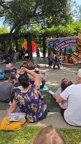 The video shows a crowd of people, mostly adults with young children, sitting on steps outdoors, watching a performance by two musicians. The musicians, a man and a woman, are sitting on chairs on a small stage. The man is playing a guitar and the woman is playing a mandolin or similar instrument. Behind them is a banner that reads "World's Tallest Leaning Tower of Smore'za!". In front of the stage is a pile of logs, presumably for a campfire. The crowd is diverse, and some people are clapping along to the music. The setting is a park or garden, with trees providing shade. A white canopy hangs over the seating area. The camera is held by someone sitting in the crowd, who is tapping a small stick.