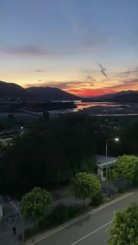 The video shows a sunset over a valley with a river. In the foreground, there is a street with a sidewalk and a few trees. A person is walking on the sidewalk. The sky is filled with warm colors of orange, red, and purple, with some clouds. The mountains in the background are silhouetted against the colorful sky. The scene is peaceful and serene.