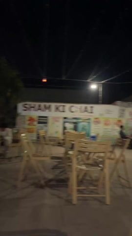 The video appears to be shot at an outdoor cafe or restaurant at night. The area is lit with strings of lights. There are several tables and chairs, some made of wood. Some people are visible in the background, either seated at tables or standing. The camera pans across the scene, focusing on two men sitting at different tables. The first man is wearing a white shirt and looking at the camera. There is a blue pitcher of water, a glass, a phone, and cigarettes on his table. The second man is wearing a white shirt and jeans. His arm is in a sling. There is a phone on his lap. He looks around the area.