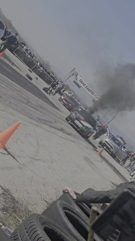 Here's a description of the video:

The video shows a drag racing event on a sunny day. A black Volkswagen Golf, heavily customized with a colorful, geometric pattern on its hood and roof, is burning out at the starting line, creating a cloud of black smoke. The car is numbered "RO.04 DUO". The "DRAG RACING" banner is visible overhead. There are two other cars staged next to it. A crowd of spectators is lined up behind a barrier on the side. Orange cones mark the track. The camera is positioned near a stack of tires, with a person partially visible in the foreground, holding onto what appears to be wooden sticks. The ground is asphalt with tire marks. There are mountains visible in the distance under a pale blue sky.