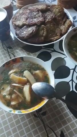This video captures a meal being prepared or served, likely a traditional Moldovan dish. The scene is set on a table covered with a patterned tablecloth featuring stylized leaves.

In the foreground, a bowl of soup is prominently displayed. The soup appears to be hearty, with visible chunks of potato, herbs like dill or parsley, and possibly meat. A silver spoon rests in the bowl, suggesting it's ready to be eaten. To the left of the soup, a white mug filled with a light-colored liquid, possibly milk or yogurt, is visible, along with some sliced bread in the background.

Behind the soup, a plate piled high with what look like potato pancakes or fritters is centered in the frame. These are golden brown and appear to be crispy. Another bowl of soup is partially visible to the right, suggesting this is a meal for multiple people.

The lighting suggests it could be daytime, possibly late morning or early afternoon. There are no indications of specific weather conditions or strong emotional cues, beyond the simple presentation of a home-cooked meal. The overall impression is one of a comforting, rustic meal. The specific location context provided (Ciuciulea, Moldova) further reinforces the likelihood of this being a traditional Moldovan culinary experience.