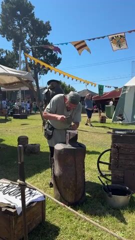 A blacksmith is demonstrating his craft at a fair. He is wearing a cap and a medieval-style tunic and trousers. He is bent over an anvil, hammering a piece of metal. In the foreground, there is a wooden chest filled with tools. In the background, there are tents, flags, and people. The sky is blue and the sun is shining. The event appears to be a historical reenactment or renaissance fair.
