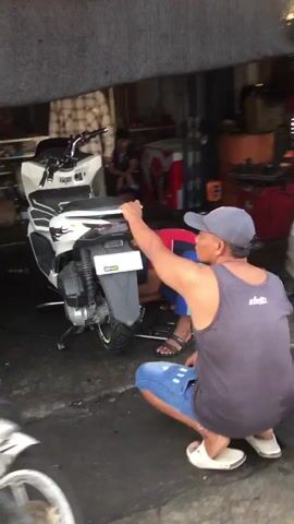 The video shows two men working on a white and black scooter in what appears to be a workshop or garage. One man, wearing a grey tank top and blue shorts, is squatting behind the scooter and appears to be adjusting something near the rear fender. He is wearing a baseball cap. The second man, wearing a red shirt and blue pants, is crouched down in front of the scooter, also working on it. The scooter has a license plate and is positioned on a stand. The environment is dimly lit and cluttered with various items, including tools and equipment, suggestive of a repair shop.