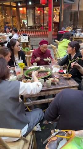 A group of six people are gathered around a wooden table outdoors, enjoying a meal in what appears to be a bustling public space. The table is laden with various dishes, bowls, drinks, and what seems to be a large, central hot pot. The people around the table are using chopsticks and spoons to serve themselves and eat. One elderly woman, wearing a red beret, is prominently positioned at the table. The other members of the group are younger, and their clothing styles vary. The environment around the table has an outdoor dining area vibe with buildings in the background, one with Chinese characters on a red banner. People are walking around in the background. The table and chairs appear somewhat rustic and aged, adding to the overall ambiance of the scene.