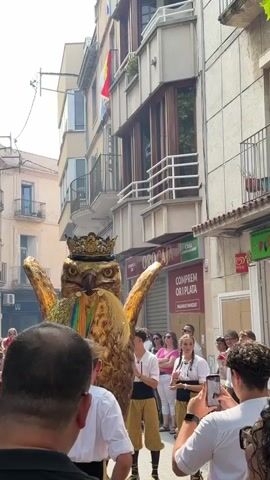 The video shows a street celebration. A large, golden eagle costume with a crown is the central figure. The eagle has its wings spread and is being carried or moved by people. In the foreground, several people are looking at the eagle, some with phones out to take pictures. Others are dressed in white shirts and some sort of tan pants or leggings. In the background, there are buildings with balconies and flags, suggesting a European setting. A Spanish flag is visible on one of the balconies. There are also shops and businesses lining the street. The overall atmosphere appears festive.