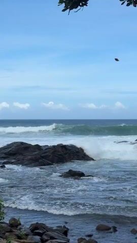 A natural ocean scene shows waves crashing against dark rocks. The sky is blue with scattered white clouds. A bird is visible in the sky. The water is choppy and appears to be dark blue. The overall scene is serene and natural.