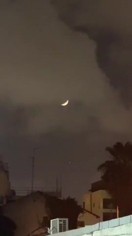 A nighttime shot shows a crescent moon in a cloudy sky above a city. The moon is bright yellow against the dark gray clouds.  Below the moon, a faint dot of light is visible. Silhouetted rooftops, antennas, and a palm tree are in the foreground.  An air conditioning unit sits on a roof. The scene is still.