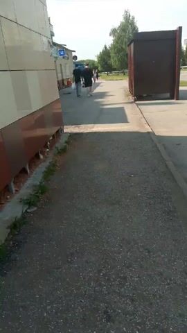 A video shows a sidewalk or path next to a building. Two men are walking away from the camera down the path. The building on the left has large beige and brown panels. The path is made of asphalt. In the background, there are more buildings, trees, and a clear sky. The lighting suggests it is daytime, likely sunny.