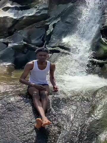 A young man with short dark hair sits on a rock facing the camera, against a backdrop of a small waterfall cascading down a rocky cliff. He wears a white tank top and dark swimming shorts, and is barefoot.  He is wet, and is splashing water at the camera. The environment is rocky and wet, with greenery visible on some of the rocks.