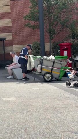 A man wearing a blue shirt, navy shorts, a blue cap, and a high-visibility vest is standing next to a trailer filled with gray bins and green bins. The trailer has two wheels and appears to be a mobile waste or recycling unit. To the left of the man, a woman with gray hair is sitting on a white bench, looking at her phone. In the background, a red post box and some greenery are visible. To the right of the trailer, a woman in a floral dress is pushing a stroller with a child inside. The scene takes place on a paved outdoor area with a brick building in the background. The sun is shining, casting shadows.