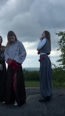 The video shows four people, seemingly young women, standing on a rocky outcrop overlooking a green landscape under a cloudy sky. On the left, a person in white pants, a patterned green top, and a large red scarf is partially visible. Next to them, a woman in a light-colored shirt with wooden toggle closures and a dark brown skirt is looking down. To the right, another woman is wearing a grey vest over a white long-sleeved shirt and a grey pleated skirt with floral accents at the waist. She is looking upwards and to the right, with her arm raised. The fourth person, mostly out of frame on the far right, is facing away from the camera. The person in the center holds a red, open box-like object. The environment appears natural and somewhat wild, with trees and distant hills visible. The lighting suggests it is late afternoon or early evening.