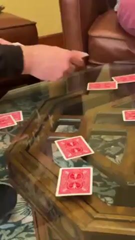 A person's hands place red-backed playing cards face-down on a glass-topped coffee table. Several cards are already arranged on the table. The table has a dark wood frame with a repeating pattern of square openings. Part of a brown leather sofa is visible in the background, along with someone wearing pink pants seated on the sofa. A small brown side table or footstool sits to the left of the sofa.