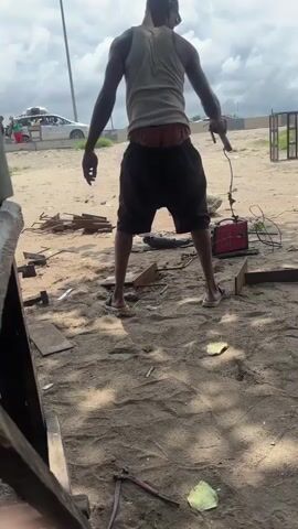 A low-angle shot captures a man standing outdoors in what appears to be a sandy workshop or construction area in Akodo, Nigeria. The man, viewed from behind, wears a white tank top, dark shorts, and sandals, with the waistband of his patterned underwear visible. His arms are slightly bent, and he seems to be observing his surroundings or waiting.

The ground is sandy with patches of shade. Scattered around are various metal pieces, tools including a pair of pliers and a hammer-like object, and a green leaf. To the man's right, a red welding machine is visible with wires connected. In the background, a white car is parked, and other people are partially visible. A metal gate frame stands further back. The sky is partly cloudy. The prominent sound of an engine or machinery can be heard throughout the video, suggesting ongoing work in the vicinity.