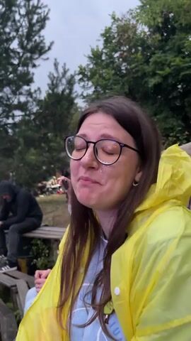 A young woman with long brown hair and glasses is wearing a bright yellow raincoat over a light blue t-shirt. She is smiling broadly and looking up and to her right. Her mouth is open, and she appears to be laughing. She has glitter on her face. In the background, other people are visible sitting on wooden benches and standing. Trees and foliage are present in the environment. The lighting suggests it is daytime, possibly overcast.