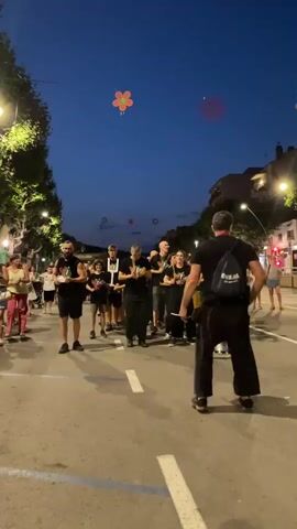 A group of people are on a street at dusk, some holding drumsticks. They appear to be part of a performance or parade. Several flower-shaped lanterns are visible in the sky. One person, facing away from the camera, is wearing a black shirt and backpack. The street is paved with white lines. Trees and buildings are visible along the sides of the street.