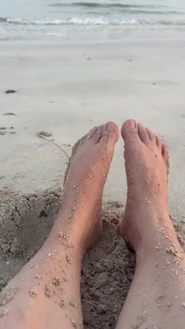 A first-person perspective shot of a person's feet covered in sand on a beach. The camera is positioned low, looking upwards towards the person's feet and legs. The sand is light brown and somewhat damp, clinging to the skin. The toes are clearly visible and appear slightly spread apart. Beyond the feet, a sandy beach stretches out, leading to a calm ocean with gentle waves rolling towards the shore. The sky is overcast or hazy, casting a soft light on the scene. The overall impression is one of relaxation and a peaceful beach setting.