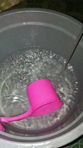 A pink plastic dipper rests at the bottom of a large grey plastic basin filled with clear water. A stream of water pours into the basin from the upper right corner of the frame, creating ripples on the surface. The water level is gradually rising. The basin is situated on a tiled floor.